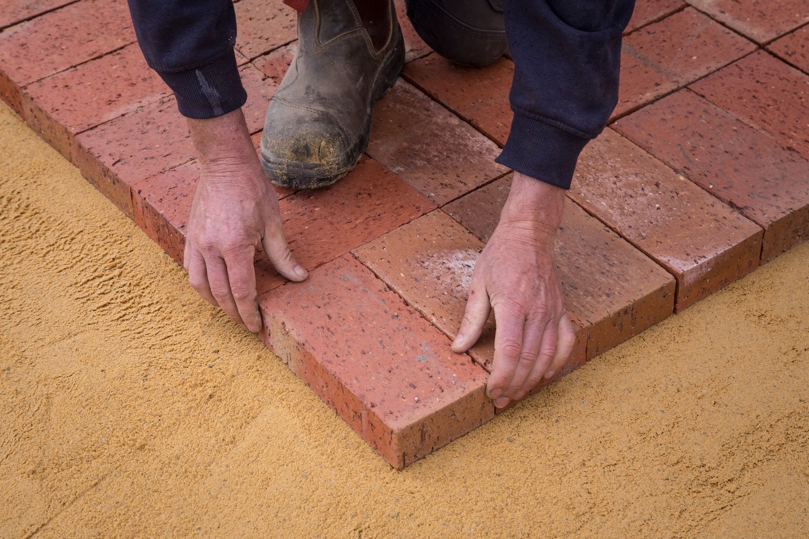 A worker sets brick pavers into a sand bed for a new patio